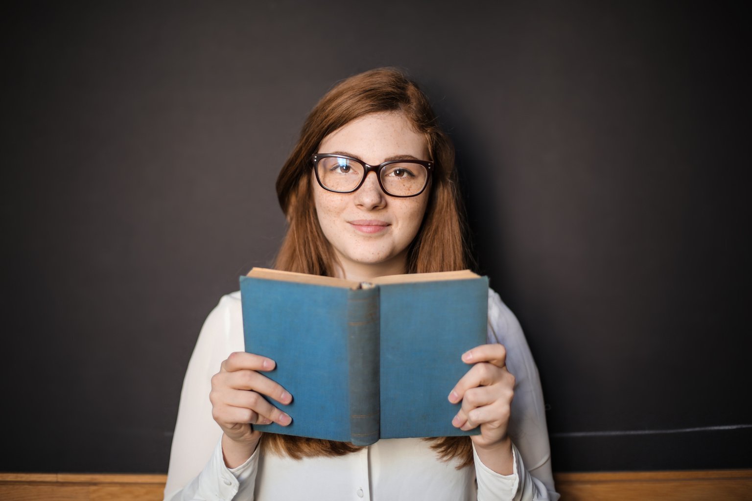 Woman in White Long Sleeve Shirt Wearing Eyeglasses Holding Blue Book